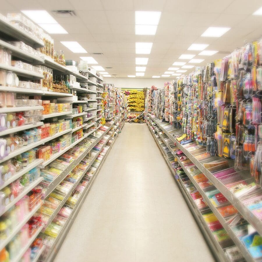 A brightly lit store aisle with fully stocked shelves on both sides, displaying a variety of colorful items and household goods. The aisle leads to a display of yellow products at the end.