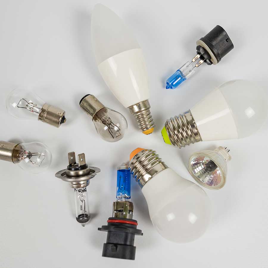 A variety of light bulbs and lamps, including LED, halogen, and incandescent types, are arranged on a white surface.