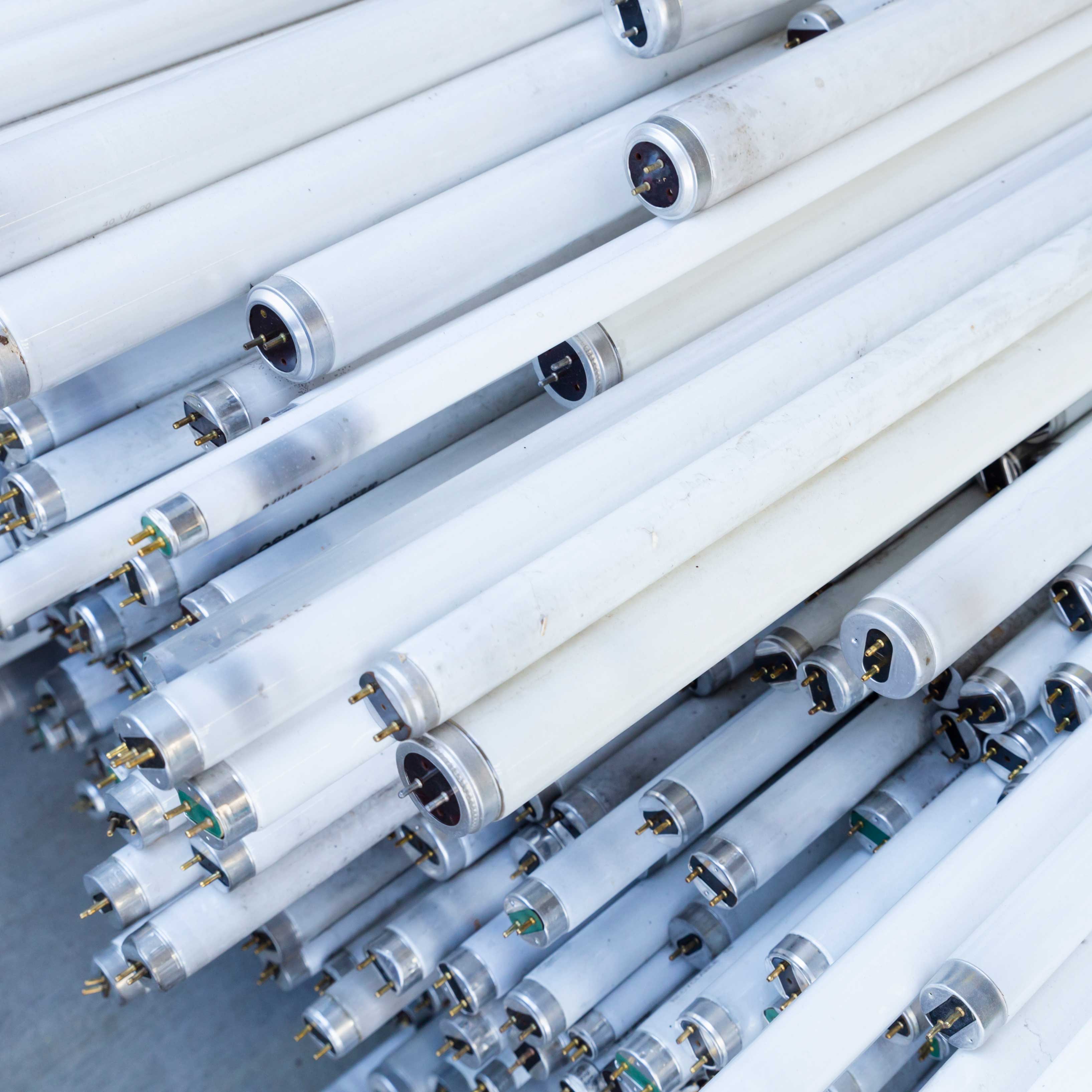 A pile of white fluorescent light tubes stacked together, showing metal end caps and prongs. The tubes are arranged in a slightly disorderly fashion, creating a pattern of repeating cylindrical shapes.