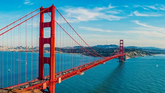 The Golden Gate Bridge in San Francisco, California, is shown spanning blue water under a bright, partly cloudy sky, with city buildings and hills in the background.