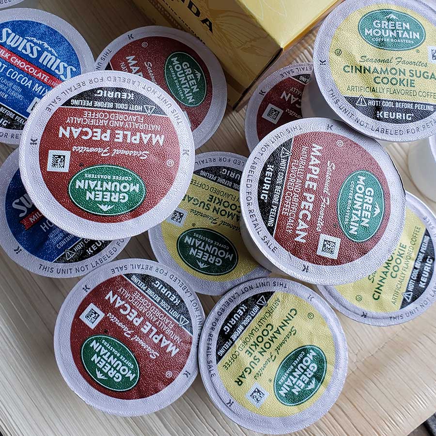 A pile of assorted coffee and hot chocolate K-Cups, including Green Mountain Maple Pecan, Cinnamon Sugar Cookie, and Swiss Miss, scattered on a wooden surface.