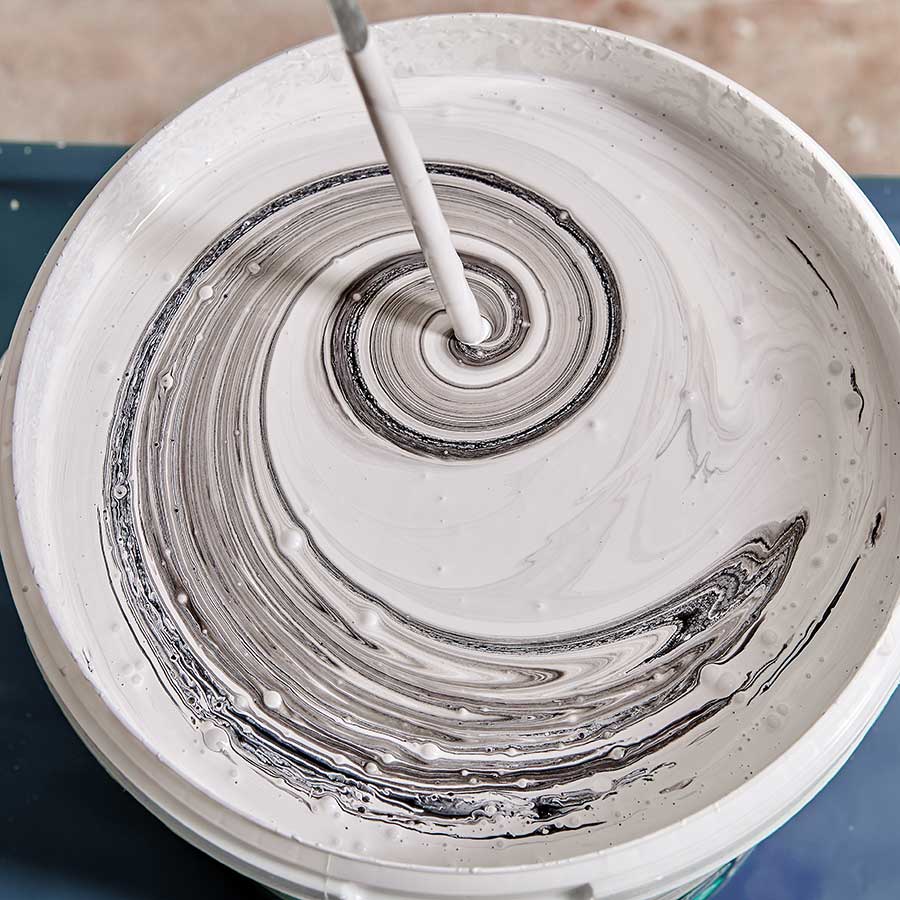 A close-up of a bucket of white paint being stirred, creating swirling gray and black patterns on the surface.