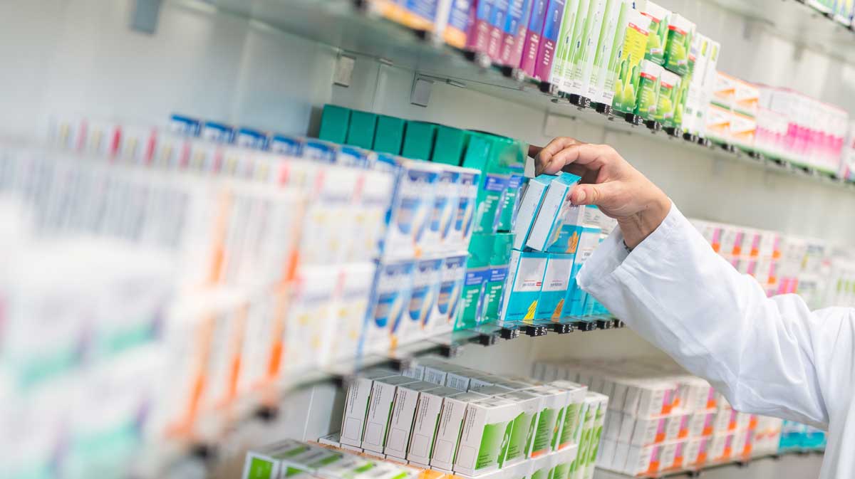 A pharmacist’s hand reaches for a box of medication on a shelf filled with various colorful medicine boxes in a pharmacy.